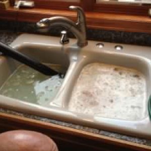 a kitchen sink filled to the brim with soapy water, a drain cleaning device is visible attempting to remove the blockage