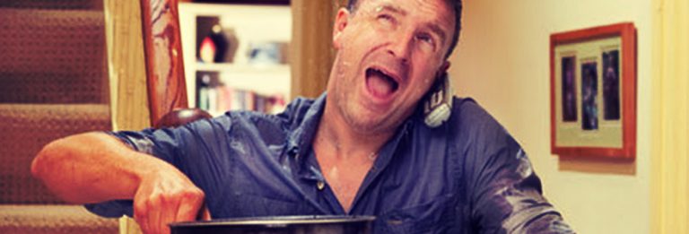 man holding buckets to catch water that is overflowing from his clogged drain in the floor above, a phone is visible in the crook of his neck as he called a plumber to help