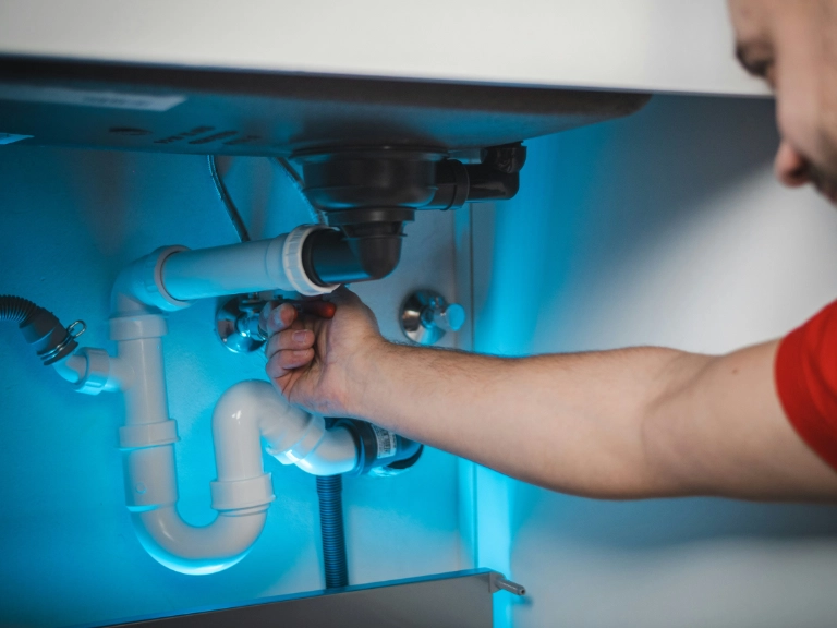 A person performs drain repair and plumbing in Toronto under a sink, tightening a white plastic pipe by hand. Various pipes and fittings are visible, while blue light illuminates the scene.