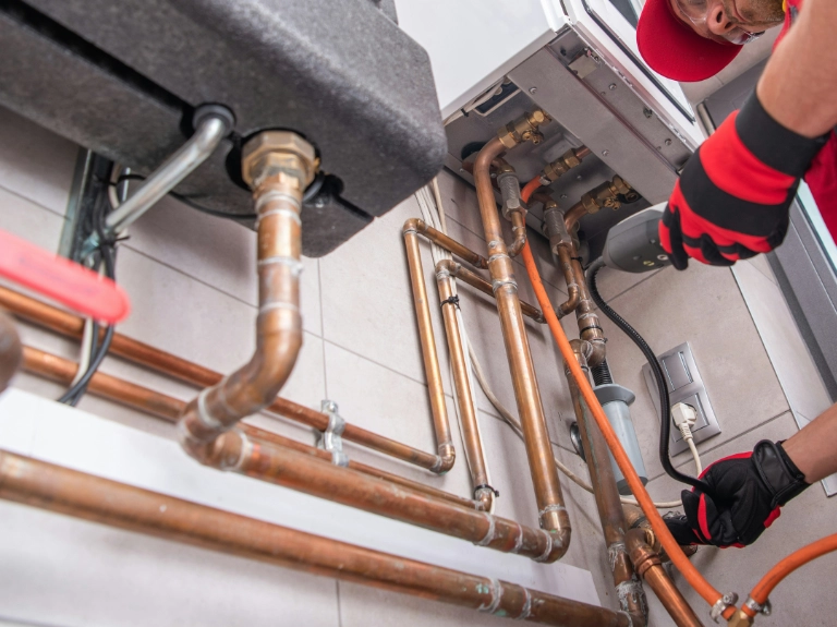 A plumber wearing red gloves and a cap is working on copper pipes and connections under a boiler in a utility room, showcasing expert drain repair and plumbing in Toronto. Various pipes, valves, and tools are visible on the tiled floor.