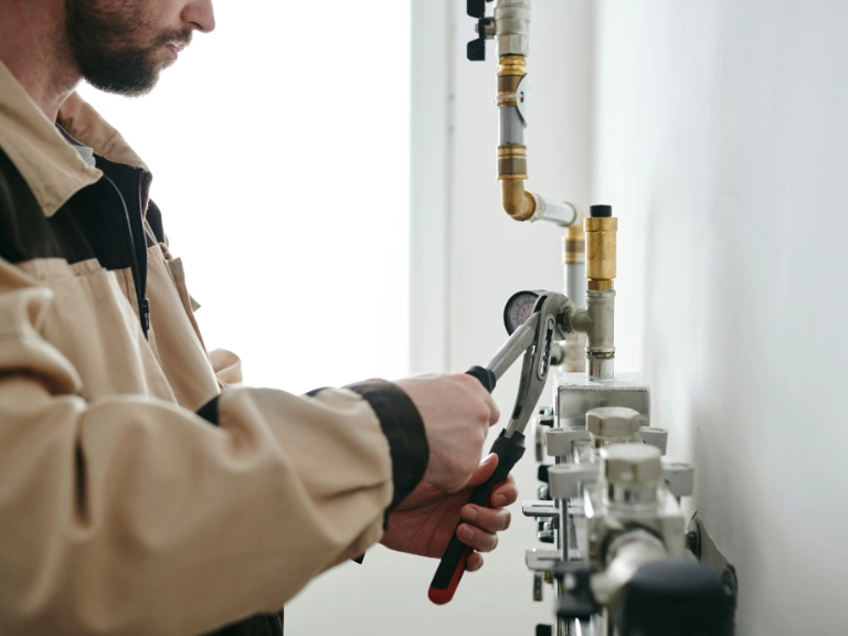 A person in a beige and brown uniform uses a wrench to adjust pipes connected to a gas or water meter on a white wall, showcasing expert plumbing in Toronto.