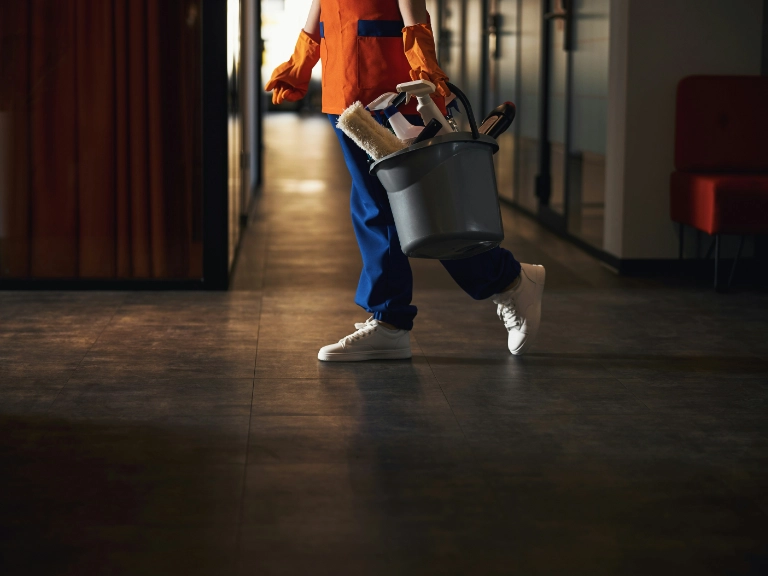 A person in cleaning attire and gloves walks down a hallway, carrying a gray bucket filled with cleaning supplies—ready for any cleaning or drain repair in Toronto. The scene is dimly lit, revealing only the lower half of the person.
