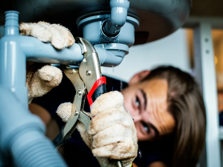 A person wearing gloves uses pliers to tighten pipes under a sink, focusing intently on drain repair and plumbing in Toronto as water leaks slightly from the pipes.