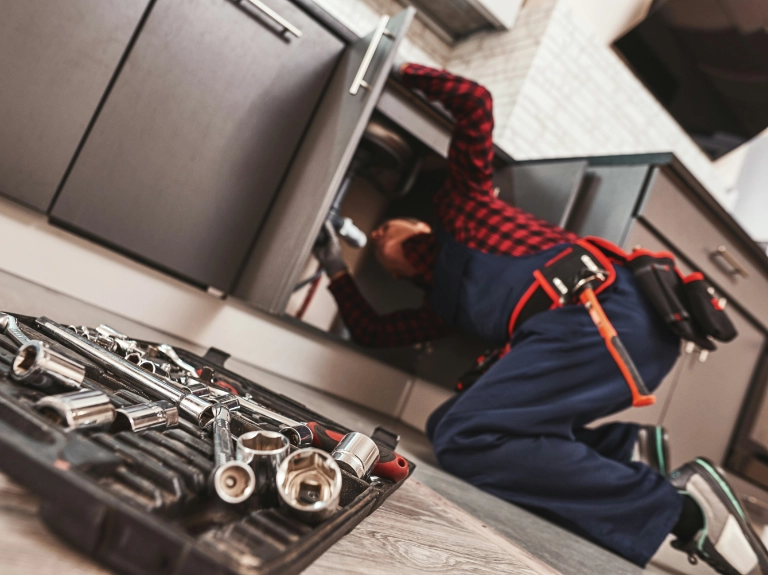 A plumber in a red plaid shirt and overalls works under a kitchen sink, surrounded by modern cabinets. An open toolbox with various socket wrenches sits on the floor, highlighting expert drain repair and plumbing in Toronto.
