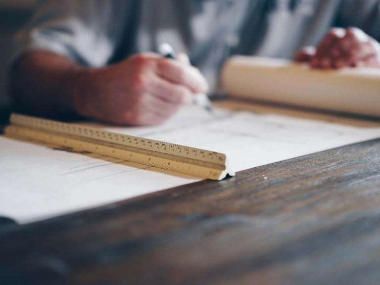 A person is drawing or writing on large sheets of paper with a pen, and a ruler is placed on the wooden table in the foreground—perhaps sketching plans for drain repair or plumbing in Toronto. The focus is on the ruler and the persons hand.