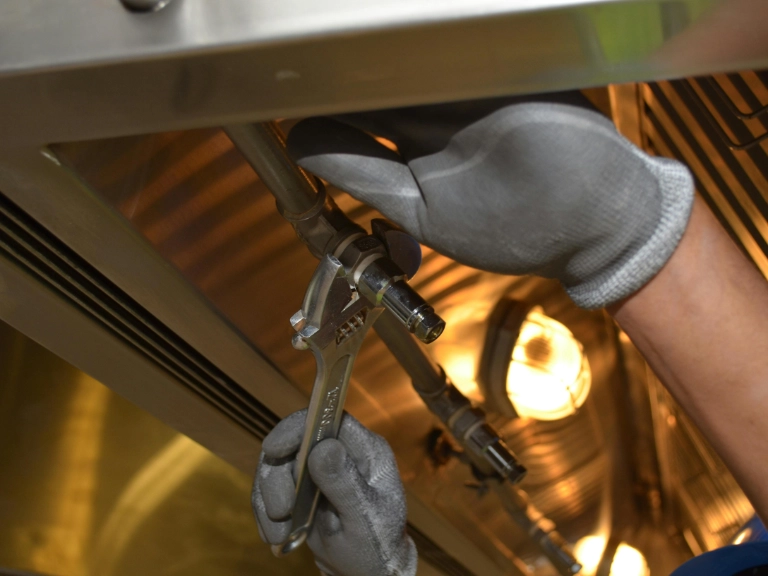A person wearing gray gloves uses a wrench to tighten a metal pipe under a metallic surface, with a light source illuminating the workspace—capturing the precision often required for drain repair and plumbing in Toronto.