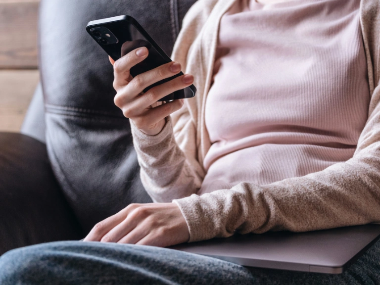A person wearing a light pink top and beige cardigan sits on a couch, researching drain repair and plumbing in Toronto on their smartphone, with a closed laptop resting on their lap.