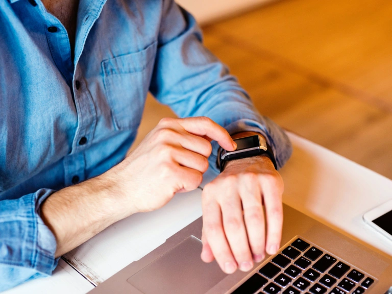 A person in a blue shirt sits at a desk using a laptop to search for drain repair and plumbing in Toronto, while checking a smartwatch on their wrist.