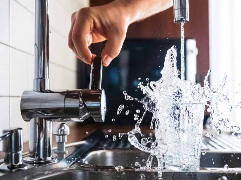 A hand adjusts the lever of a kitchen faucet while water overflows from a glass, splashing dramatically over the sink—a vivid reminder of the importance of timely drain repair and plumbing in Toronto.