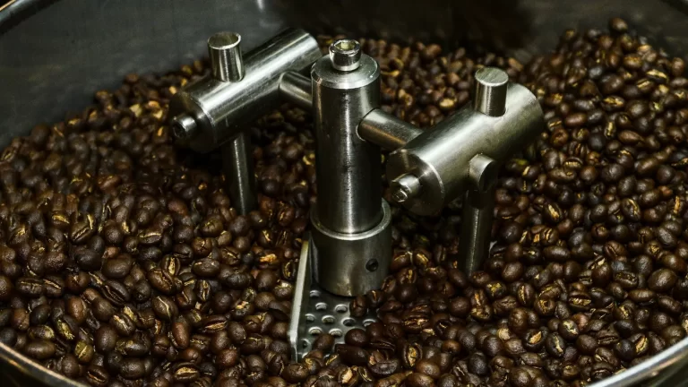 A close-up view of roasted coffee beans in a metal roasting machine, with mechanical arms mixing the beans inside the drum.