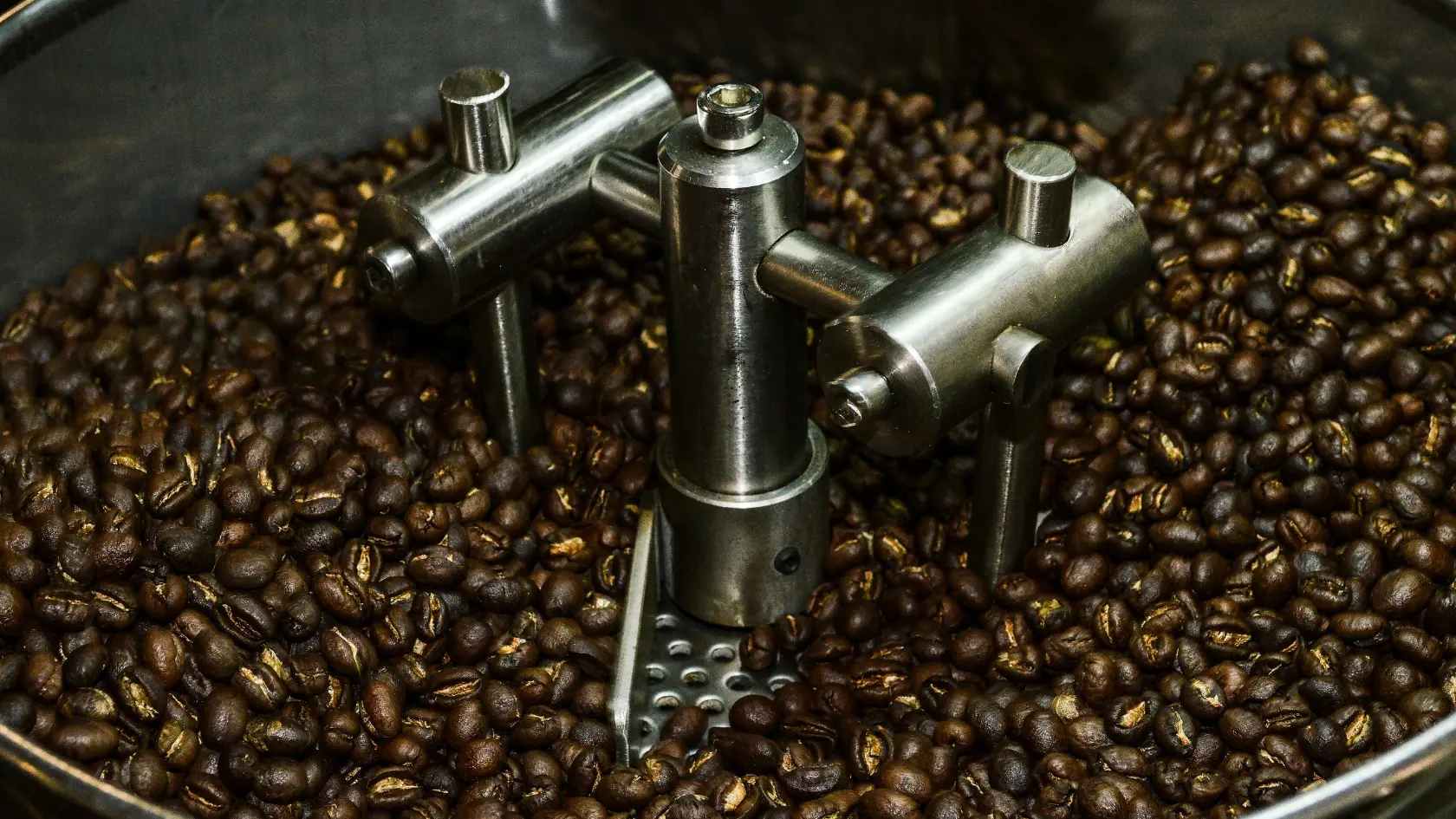 A close-up view of roasted coffee beans in a metal roasting machine, with mechanical arms mixing the beans inside the drum.