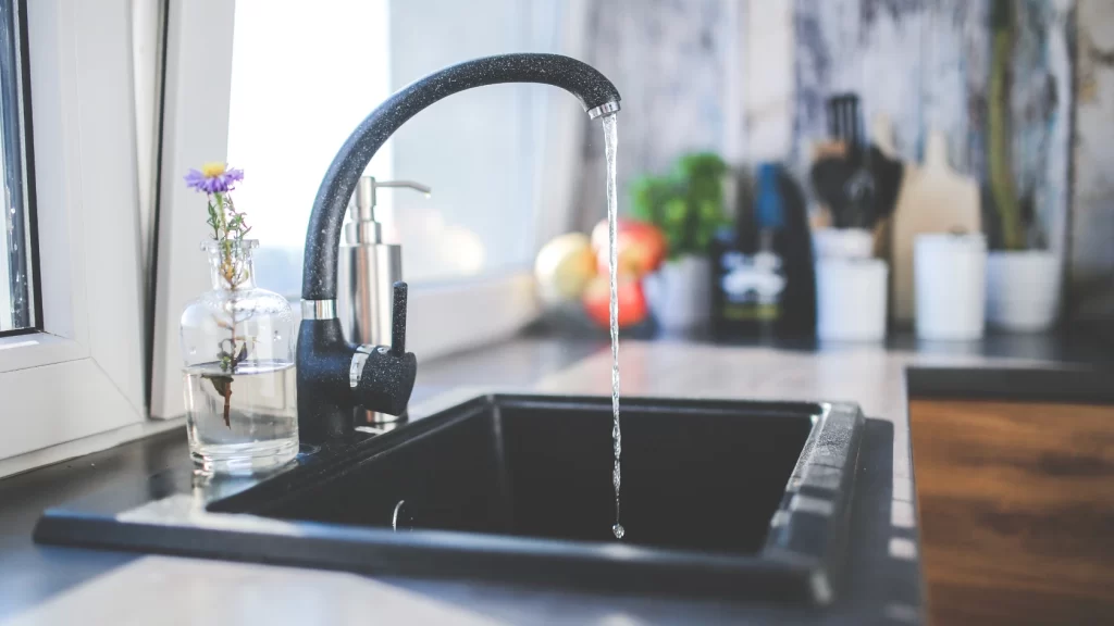 A modern kitchen sink with water flowing from a black faucet. There is a small glass vase with purple and yellow flowers and a soap dispenser on the counter, with blurred kitchen items in the background.