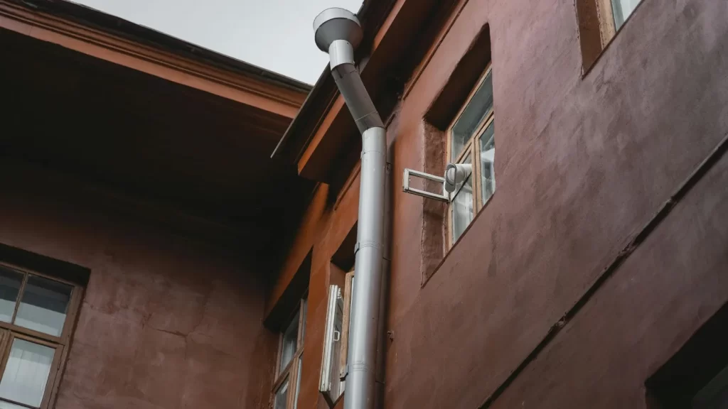 A close-up view of a building’s exterior showing brown walls, windows with white frames, and a vertical silver drainpipe running between the windows up toward the roof.