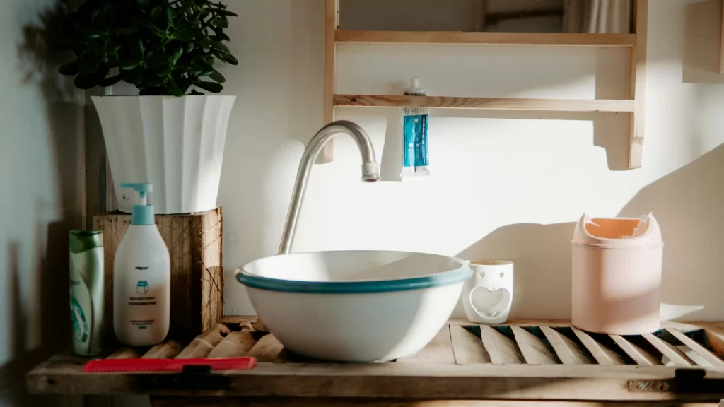 A sunlit bathroom sink with a blue-rimmed white basin, soap dispenser, potted plant, orange bin, toiletries, and a red hairbrush on a wooden countertop and shelf.