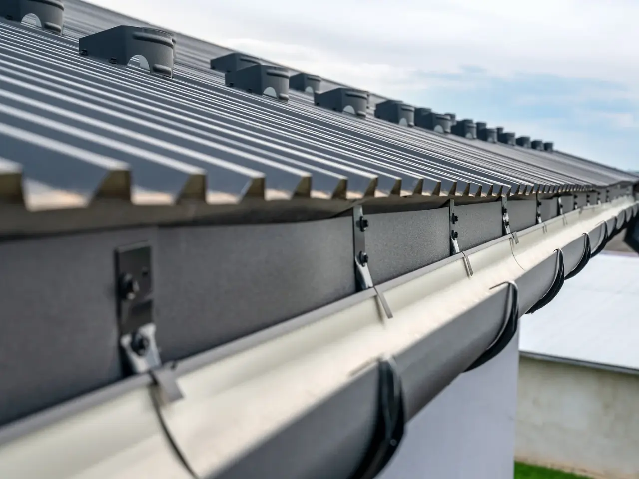 Close-up view of a modern metal roof with a row of brackets and a gutter system attached along the edge, under a cloudy sky.