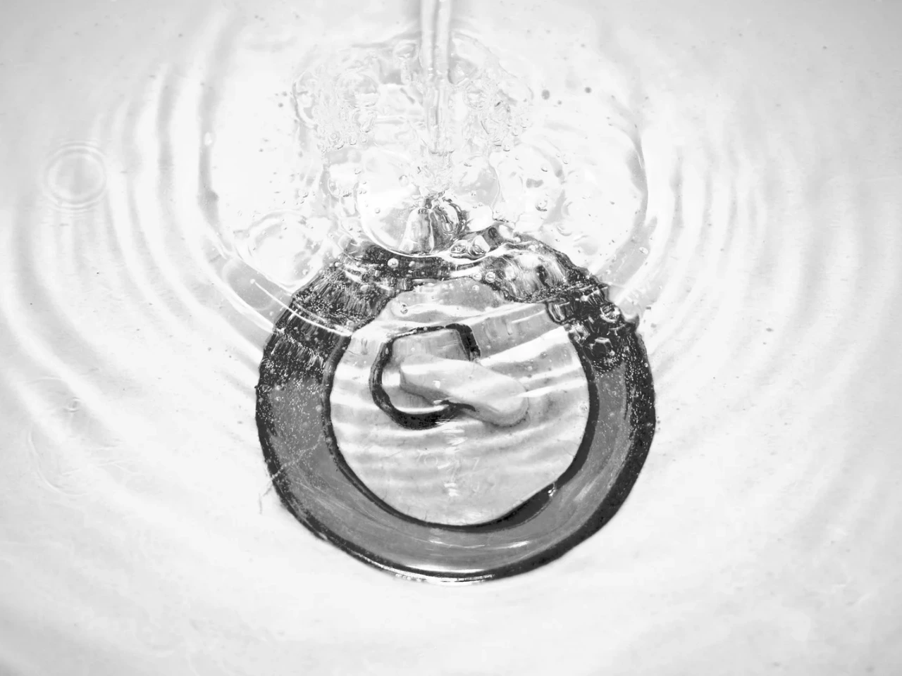 Water swirls and drains into a metal sink drain, creating ripples and bubbles on the white surface. The photo is taken from above in black and white.