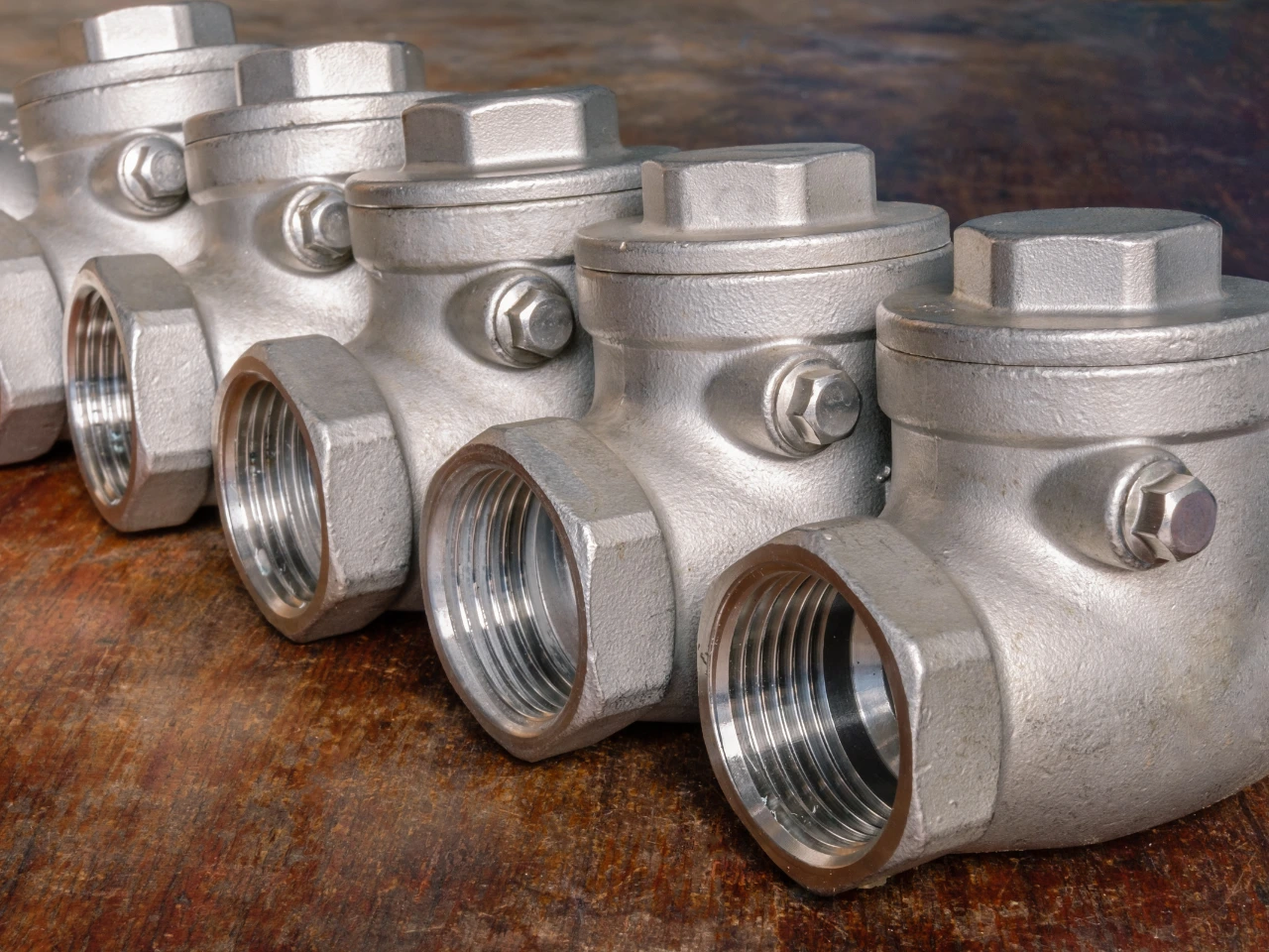 A row of metal check valves with hexagonal caps and threaded ends, aligned on a wooden surface, showing their industrial design and shiny metallic texture.