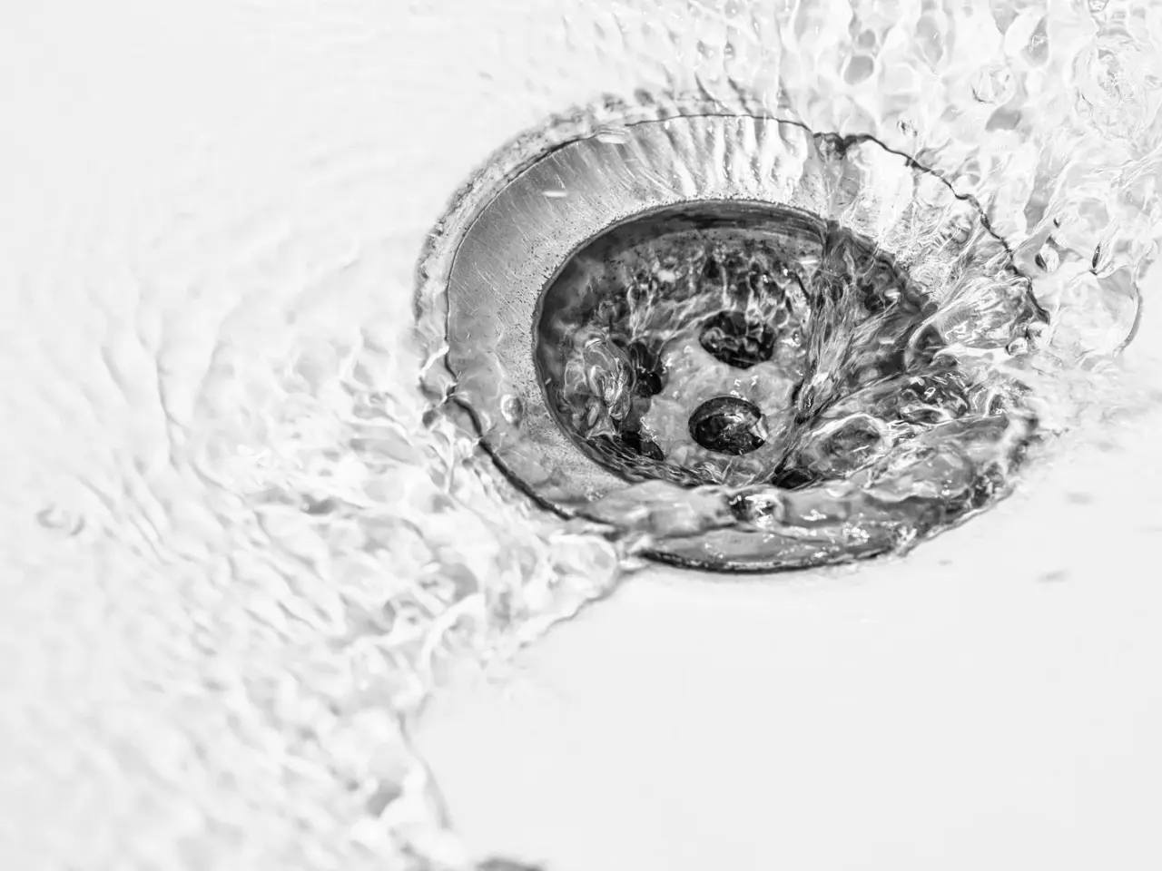 Water swirling around and draining into a circular silver sink drain, with ripples and bubbles visible on the white surface.