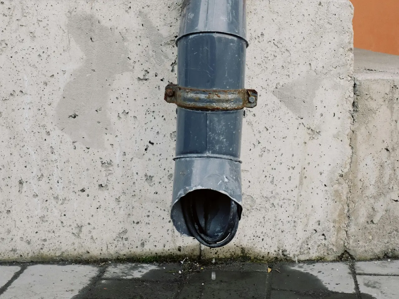 A close-up of a damaged grey metal drainpipe attached to a concrete wall, with the bottom end bent and a drop of water falling onto wet pavement below.