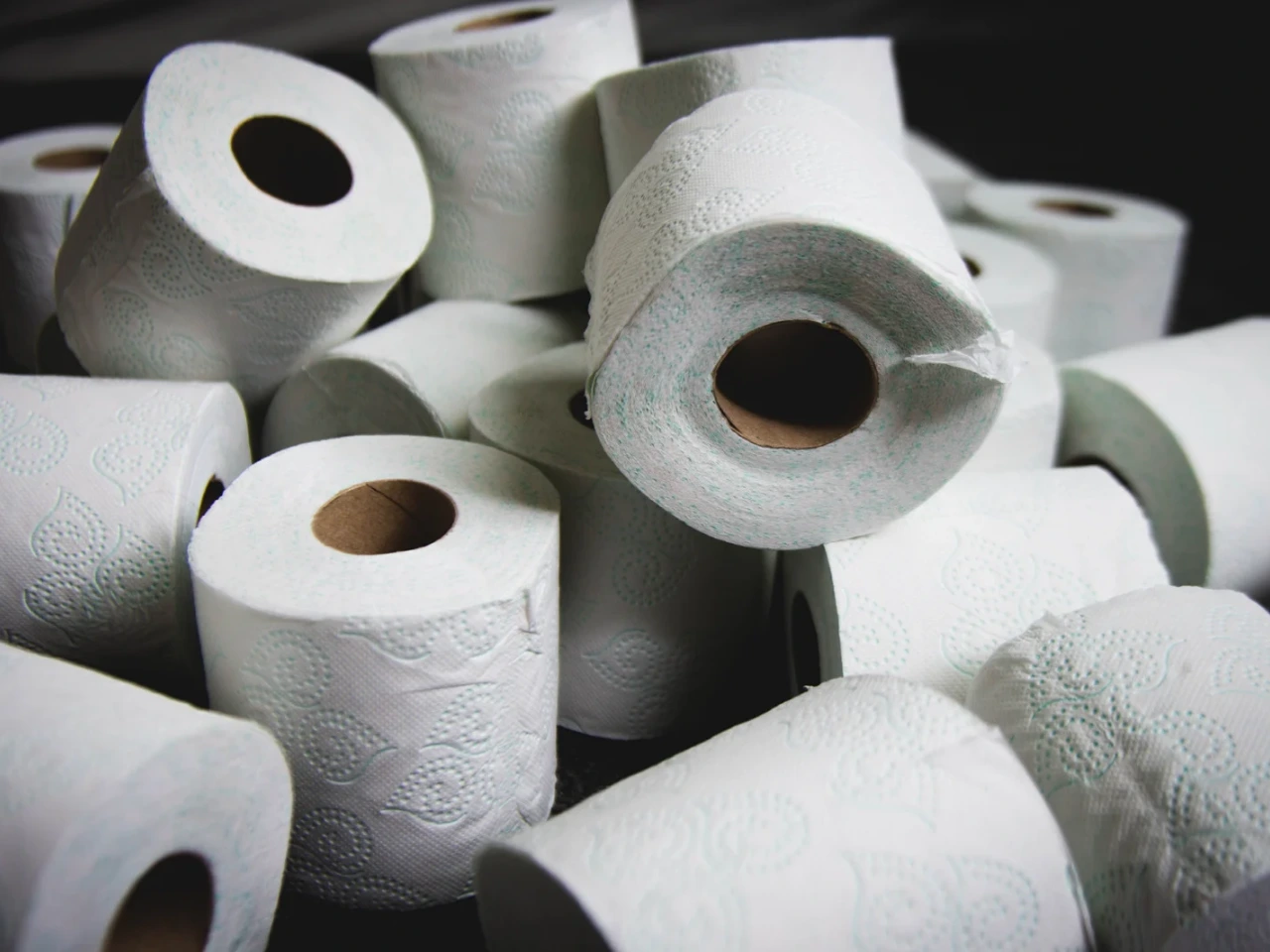 Several rolls of white toilet paper are scattered in a pile, with some rolls lying on their sides and others upright. The background is dark, making the toilet paper stand out.