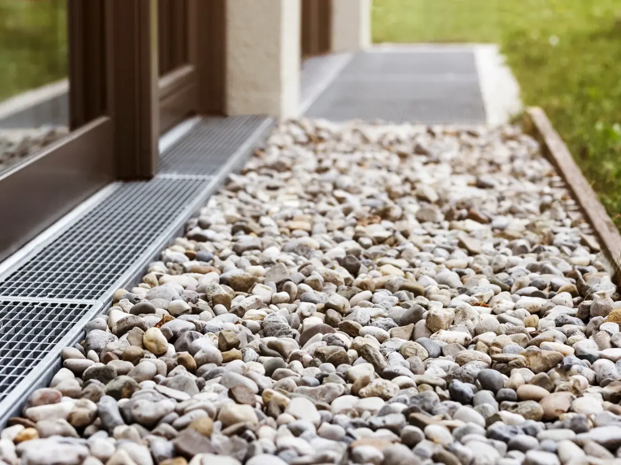 Close-up view of a gravel bed with small, smooth stones beside a building, bordered by a metal drainage grate. Grass is visible in the background.