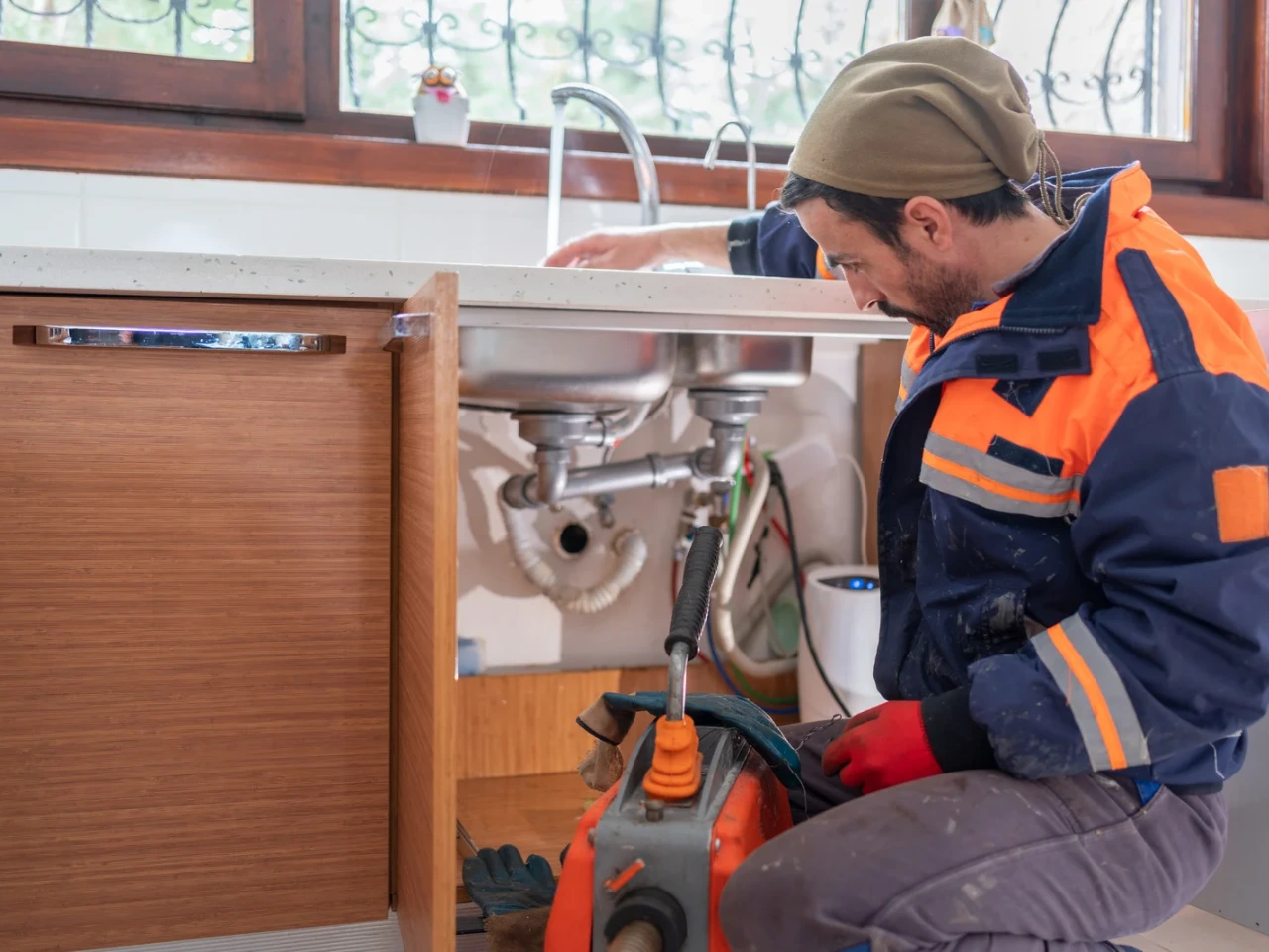 A plumber in a work uniform and beanie kneels in front of an open kitchen cabinet, working under a sink with tools and equipment, focused on repairing the plumbing.
