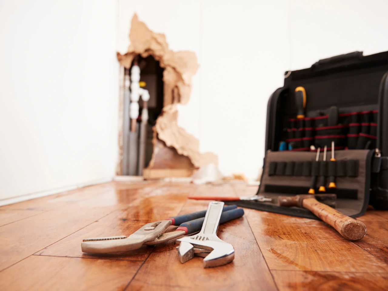 A close-up of plumbing tools and a toolbox on a wet wooden floor, with a large hole in the wall revealing pipes and insulation in the background.