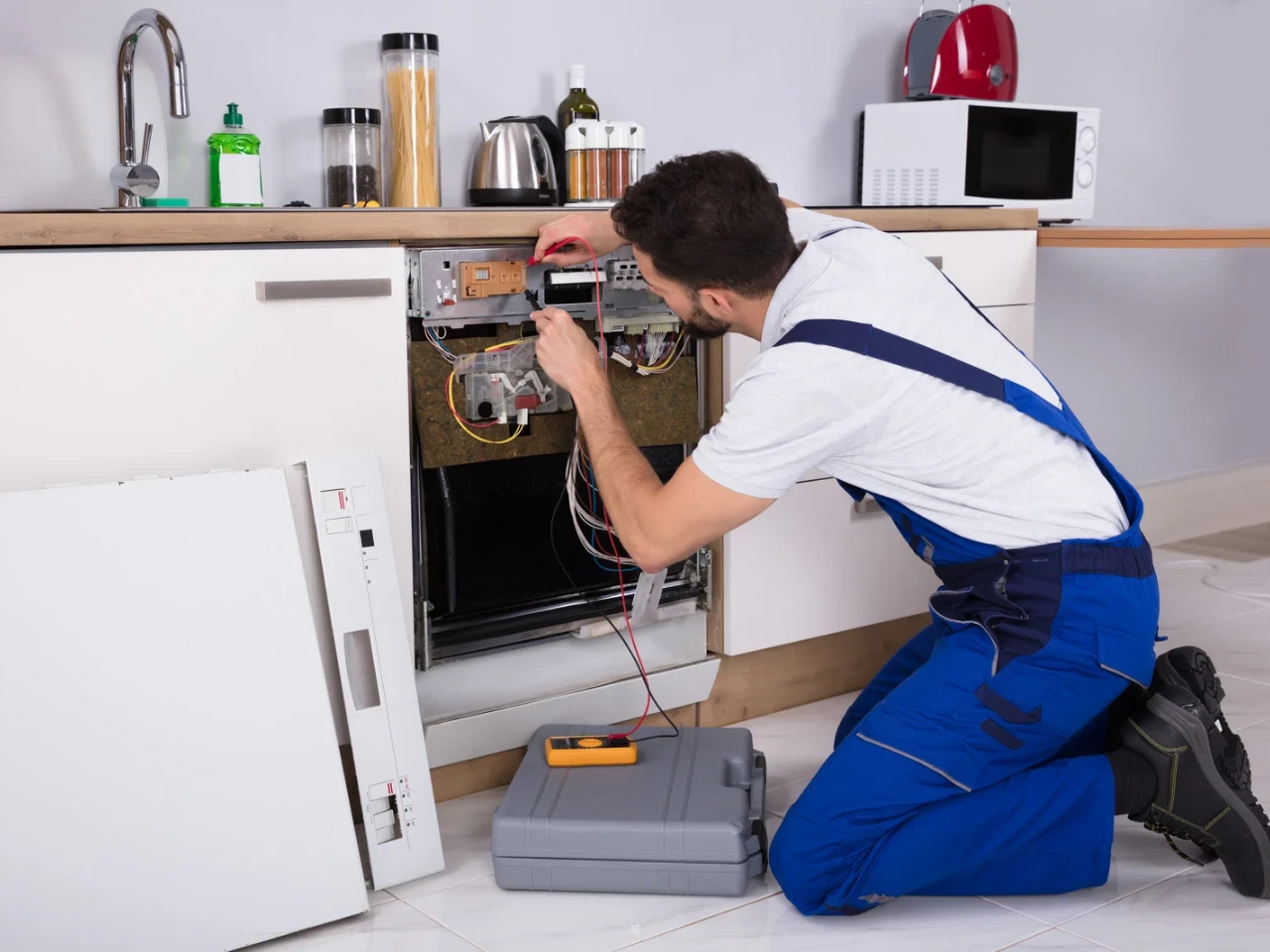 A repairman in blue overalls kneels on the floor, fixing the internal components of a built-in dishwasher with its front panel removed. Tools and a toolbox are nearby, and kitchen appliances are on the counter above.