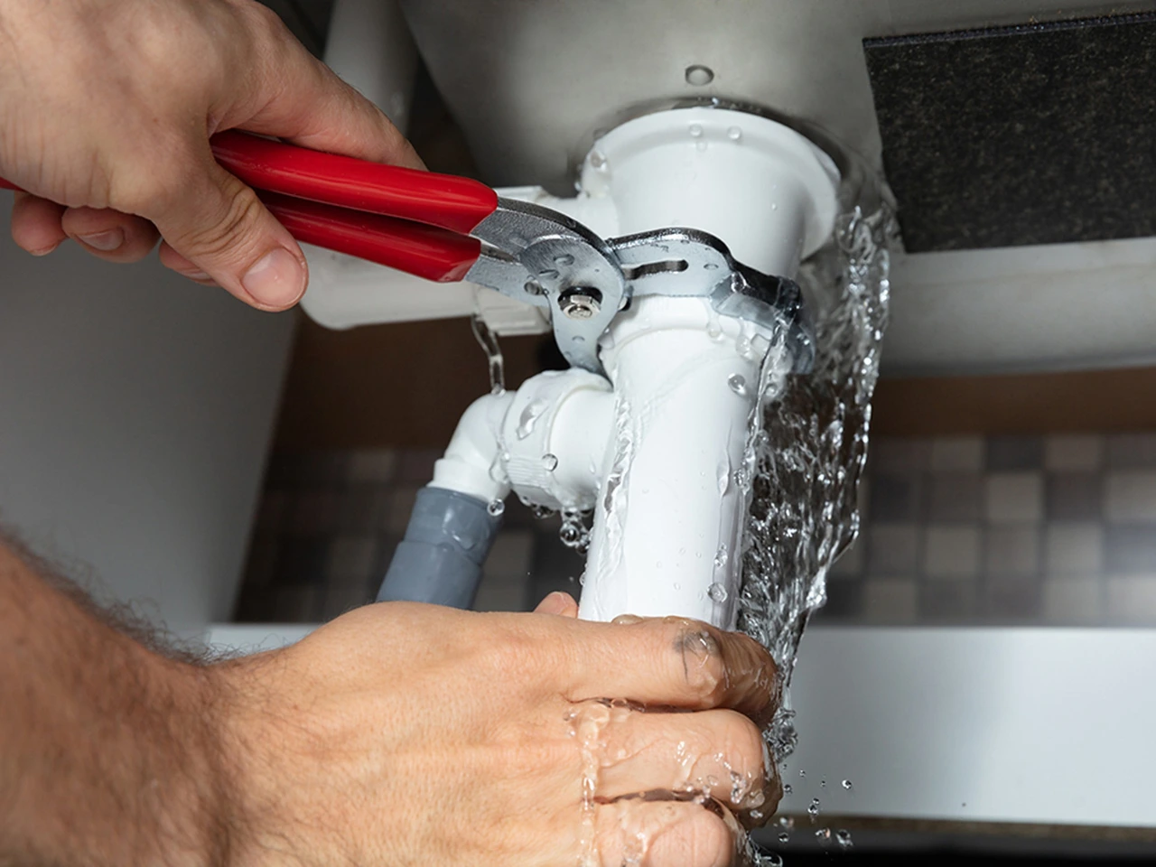 A person uses a red-handled wrench to fix a leaking white pipe under a sink, with water spilling out around the pipe connection.