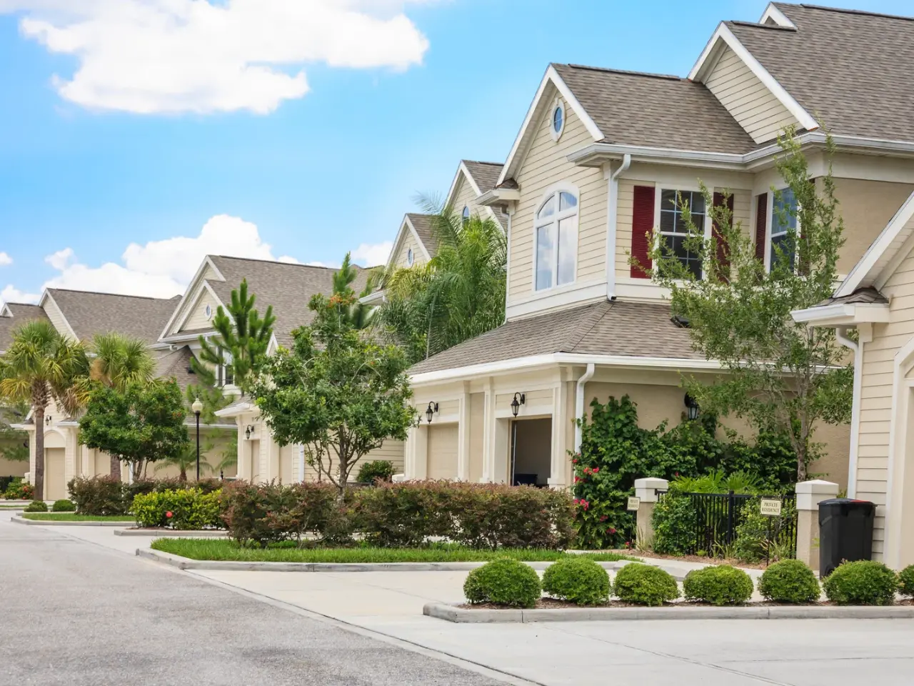 A row of suburban homes with light-colored siding, manicured lawns, green shrubs, and trees lining a clean, quiet street under a blue sky with scattered clouds.