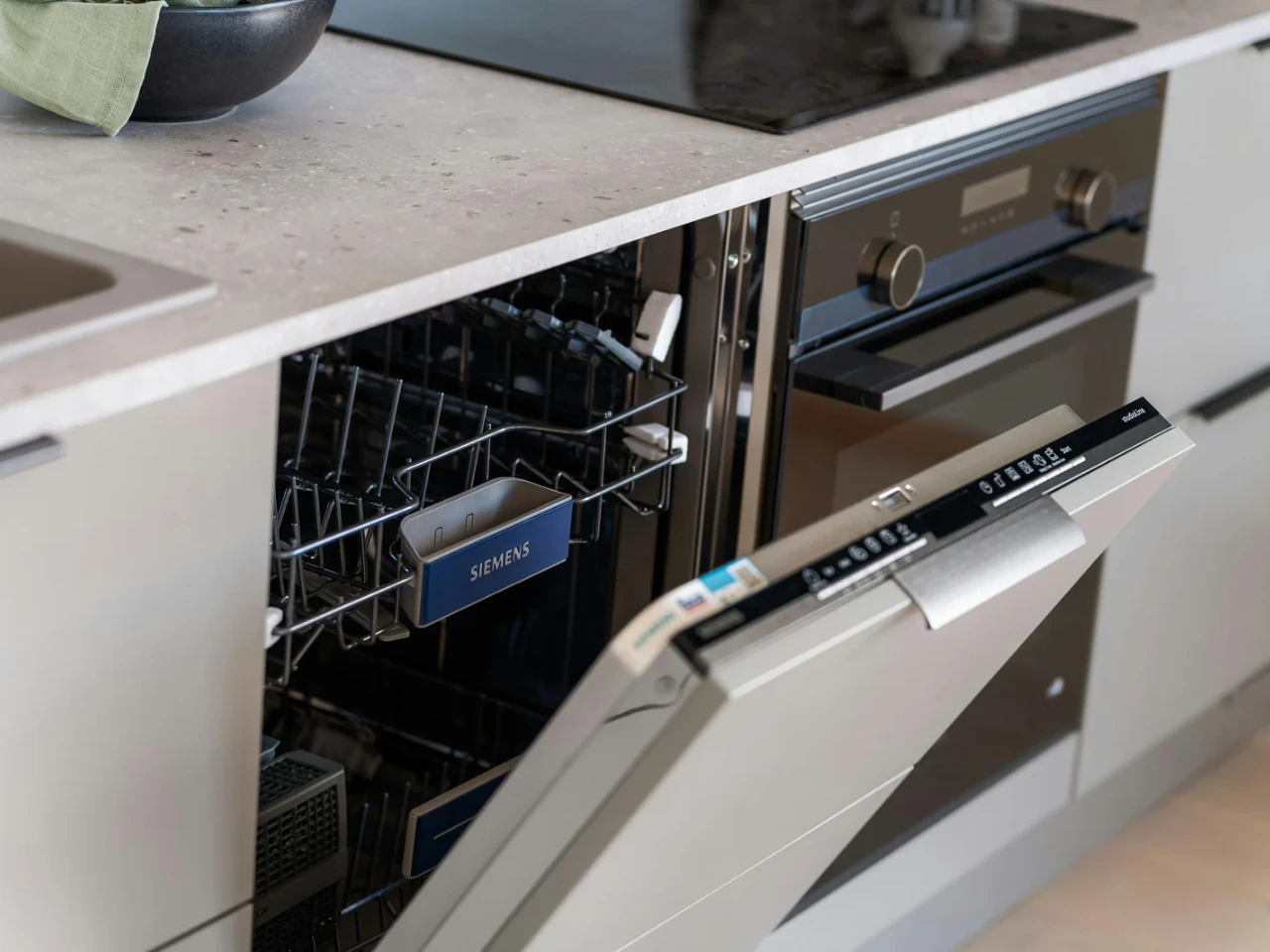 An open built-in Siemens dishwasher is shown in a modern kitchen, with the top rack partially extended. The appliances are integrated into the cabinetry beneath a light-colored countertop.