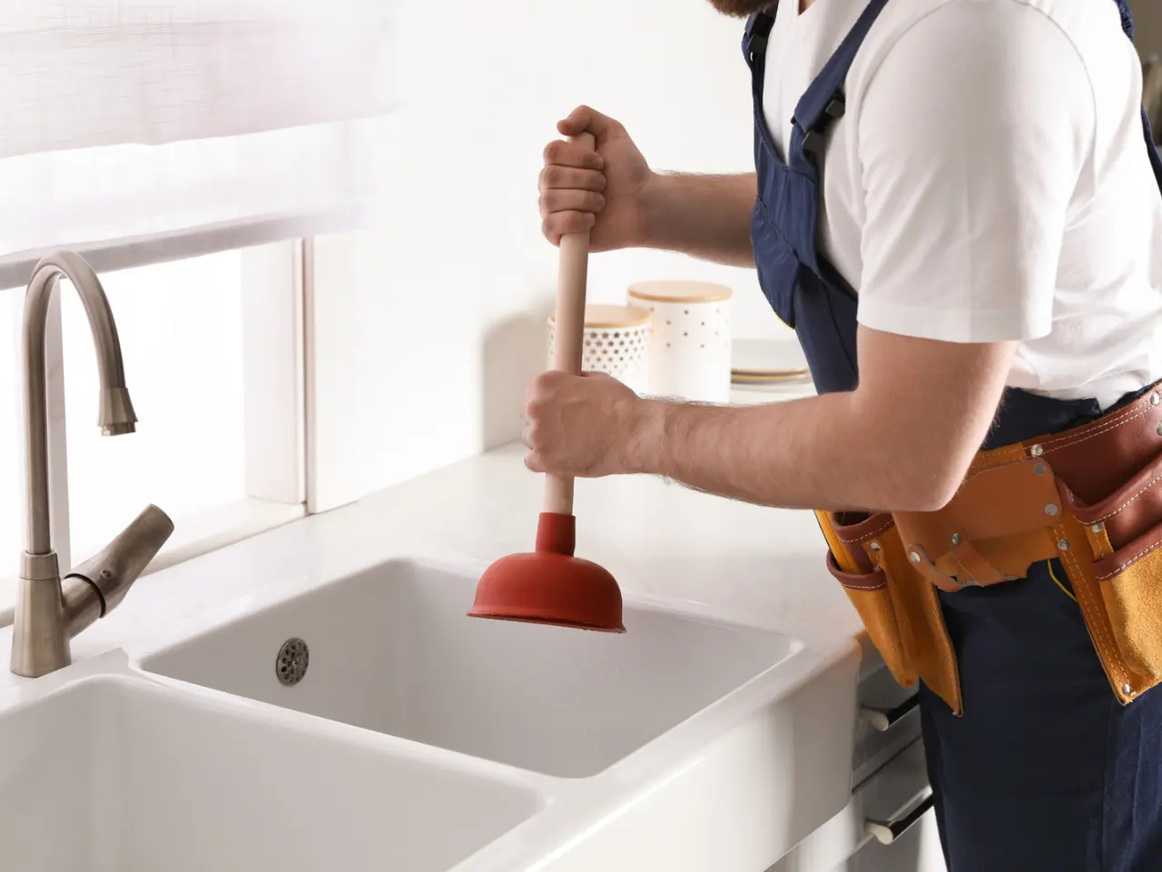 A person in a white shirt and tool belt uses a red plunger to unclog a white kitchen sink in a bright, modern kitchen.