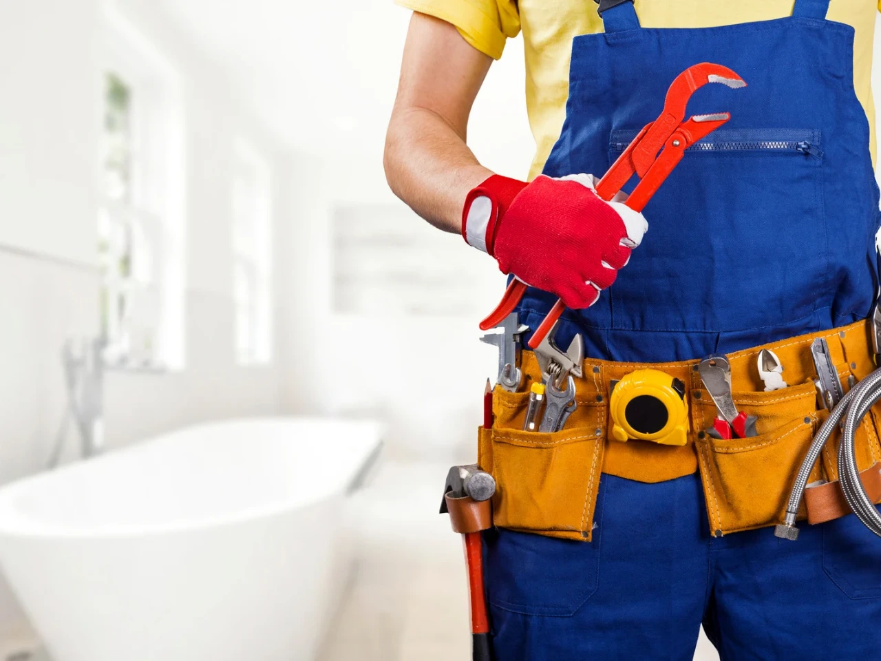 A plumber wearing blue overalls and red gloves holds a large red pipe wrench, with various tools in a yellow tool belt, standing in a bright bathroom with a bathtub in the background.
