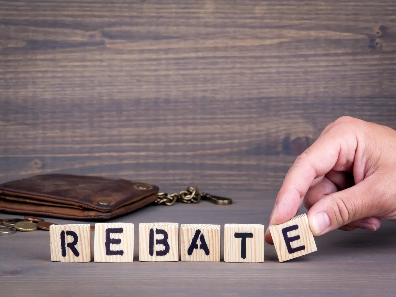 A hand arranges wooden blocks spelling “REBATE” on a table, with a brown leather wallet and chain in the background against a wooden wall.