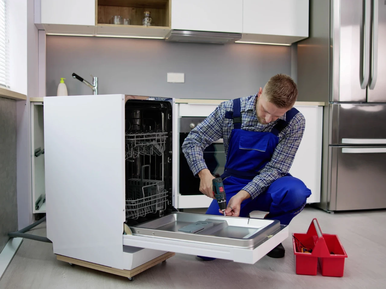 A repairman in blue overalls uses a power drill to fix the open door of a built-in dishwasher in a modern kitchen. A red toolbox sits nearby on the floor.