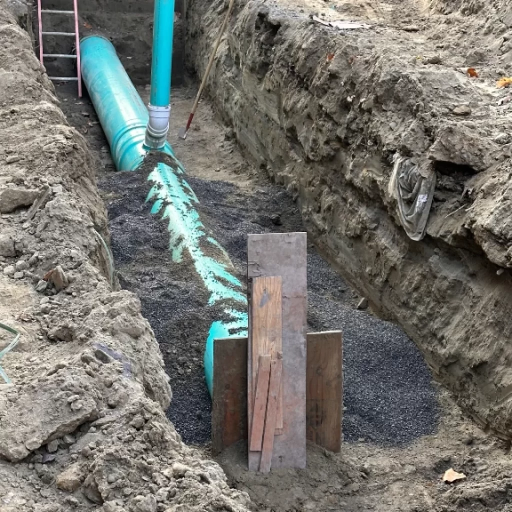 A construction trench with large blue-green pipes being installed underground for drain cleaning Etobicoke projects, surrounded by dirt walls. Black gravel encases the pipes, wooden boards stand nearby, and a ladder is visible at the far end.