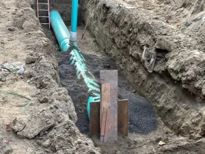 A construction trench with large blue-green pipes being installed underground for drain cleaning Etobicoke projects, surrounded by dirt walls. Black gravel encases the pipes, wooden boards stand nearby, and a ladder is visible at the far end.