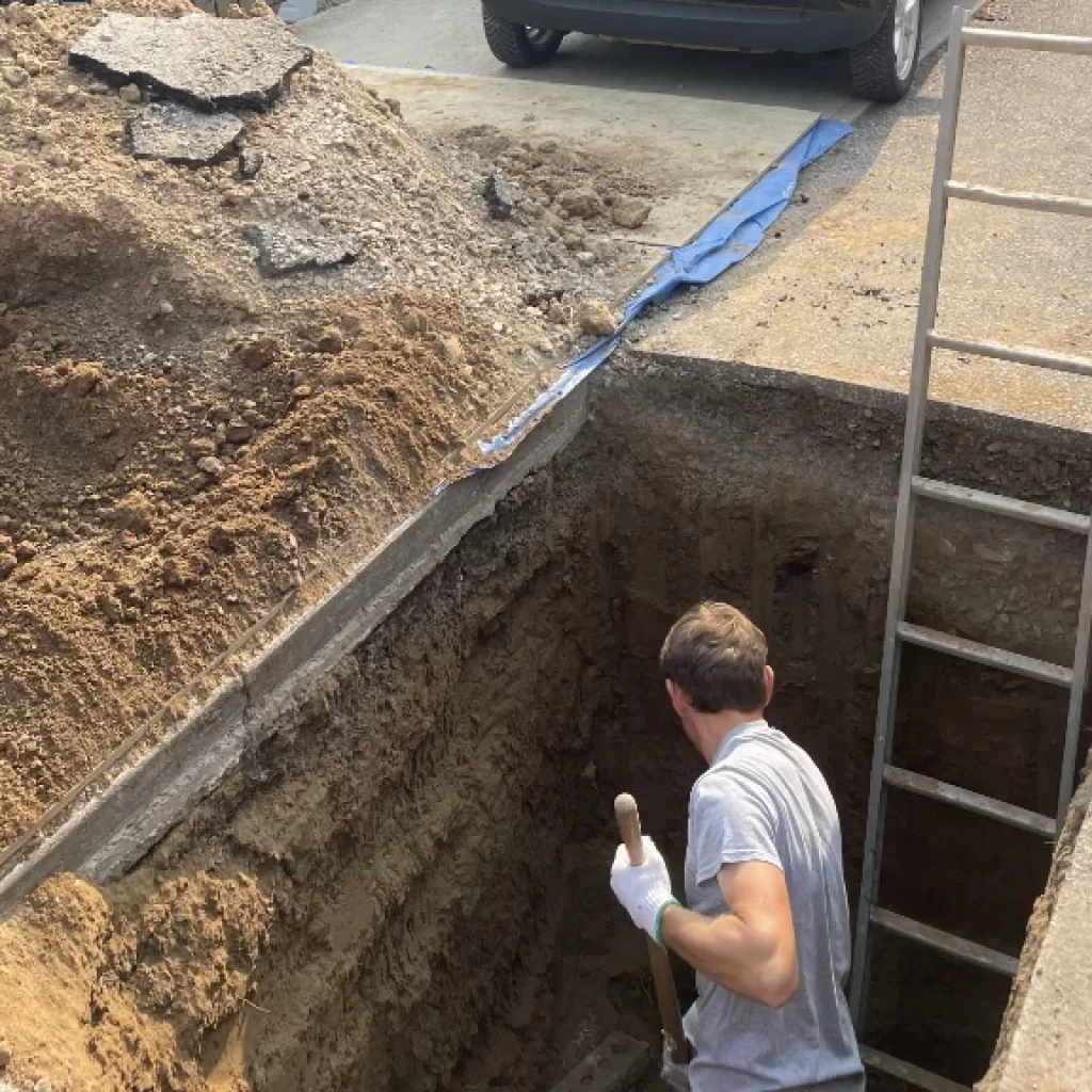 A person wearing gloves stands in a deep rectangular pit, holding a shovel, working on drain cleaning Mississauga. A metal ladder leans against the pit’s edge, with piles of dirt and broken asphalt nearby. A car is parked close to the excavation site.