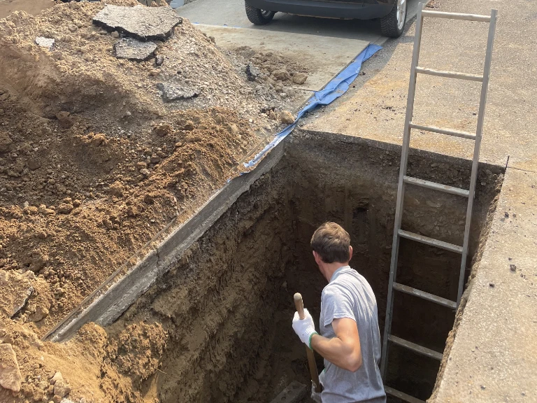 A person wearing gloves stands in a deep rectangular pit, holding a shovel, working on drain cleaning Mississauga. A metal ladder leans against the pit’s edge, with piles of dirt and broken asphalt nearby. A car is parked close to the excavation site.