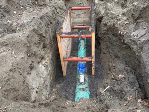 A green underground pipe exposed in a dirt trench, likely part of drain cleaning Toronto services, with a blue valve and a wooden trench box with red bars for safety. Loose soil and fallen leaves surround the work area.
