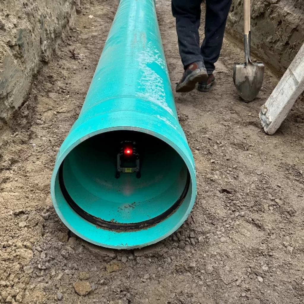 A large turquoise pipe lies on the ground at a Drain repair and Plumbing in Toronto site, with a small device emitting a red light inside. A person with a shovel stands nearby, and a ladder leans against the dirt wall.