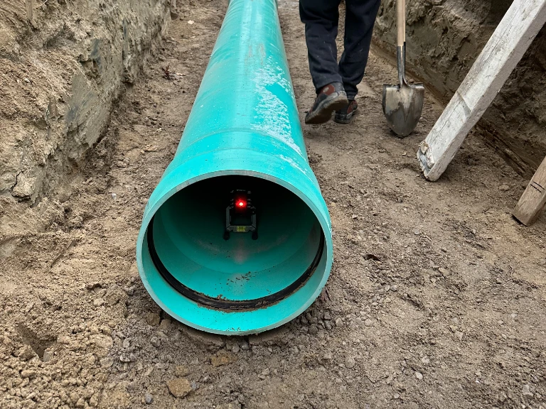 A large turquoise pipe lies on the ground at a Drain repair and Plumbing in Toronto site, with a small device emitting a red light inside. A person with a shovel stands nearby, and a ladder leans against the dirt wall.