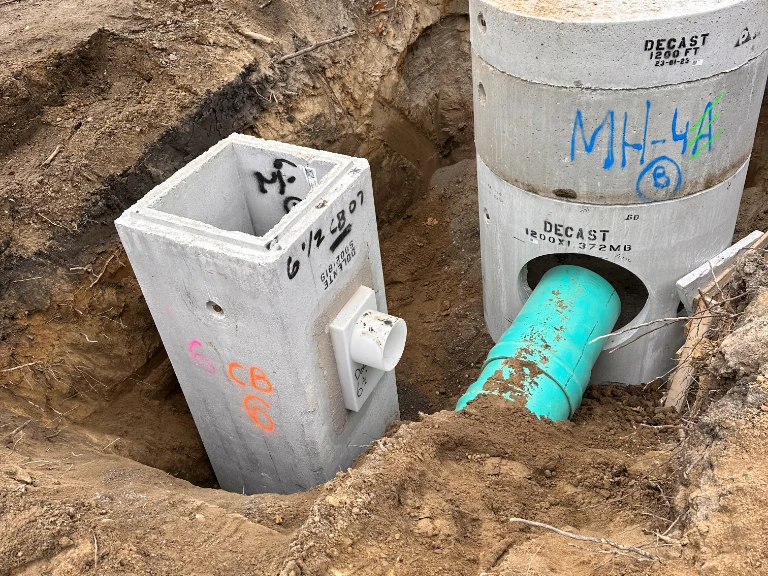 Concrete utility structures and a large green pipe, used for drain repair and plumbing in Toronto, are partially buried in a dirt trench at an excavation site, with some markings and labels visible on the structures.