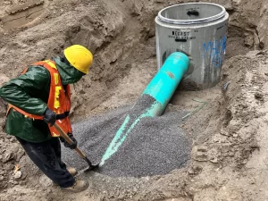 A construction worker in a yellow hard hat, green jacket, and orange vest shovels gravel around a large green pipe connected to a concrete sewer structure during drain repair and plumbing in Toronto.