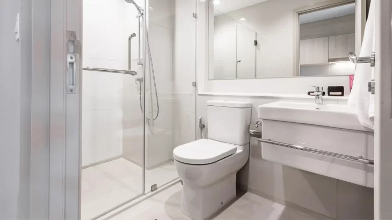 Modern bathroom with a glass-enclosed shower, white toilet, wall-mounted sink with storage, large mirror, and white walls. The space is well-lit and minimalistic in design.