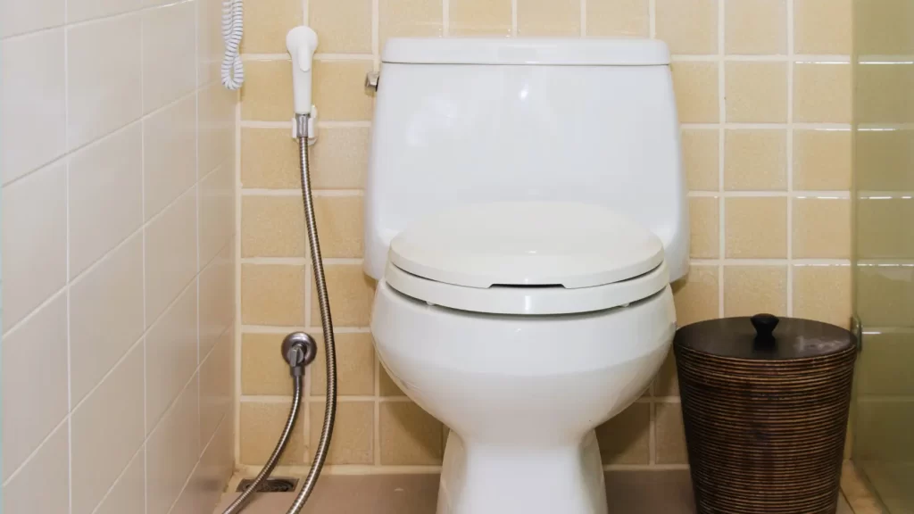 A white toilet with a closed lid sits against a wall of yellow tiles. To the left is a bidet spray attached to the wall, and to the right is a round, brown wastebasket.