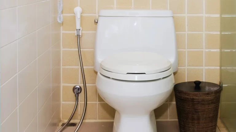 A white toilet with a closed lid sits against a wall of yellow tiles. To the left is a bidet spray attached to the wall, and to the right is a round, brown wastebasket.