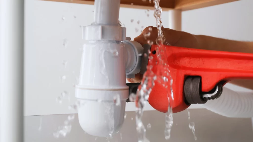 Water leaks from a white pipe under a sink as a hand uses a red pipe wrench to tighten a fitting, with water splashing around the area.