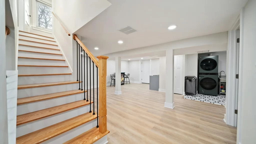 A modern finished basement with light wood flooring, white walls, a wooden staircase with black railings, a laundry area with stacked washer and dryer, and a small kitchenette or bar area in the background.