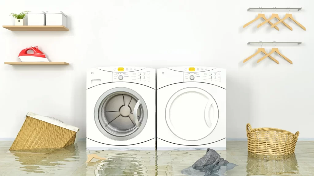 Two front-loading washing machines in a laundry room are surrounded by floodwater. Clothes, an iron, and baskets are scattered around, with some items floating in the water. Empty hangers are on the wall.